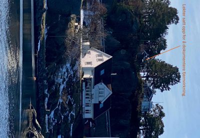 Fotografi av en hvit villa ved kysten med en annen bygning på bakken bak. Bildet inneholder en tekstmerking som peker på en bygning på bakken.