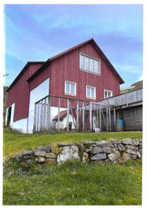 Fotografi av en rød bygning (muligens et hus eller uthus) sett fra siden, med grønt gress og en steinmur i forgrunnen.