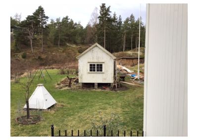 Fotografi av en liten, hvit hytte eller uthus i en hage med skog i bakgrunnen.