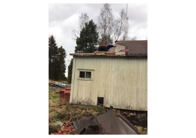 Fotografi av en hvit, paneltett bygning (kanskje en uthus eller garasje) med en person som jobber på taket. Det er også synlig med materialer på bakken og i bakgrunnen.