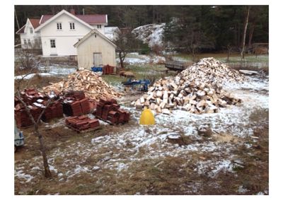 Fotografi av en byggeplass med materialer (treverk, taktegl) og eksisterende hus i bakgrunnen.