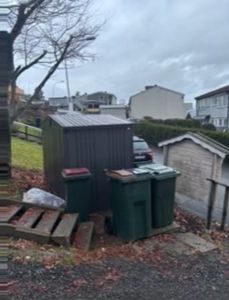 Fotografi av en tomte med et mørkt skur, to grøne søppelcontainere og en mindre hvit uthus i bakgrunnen.