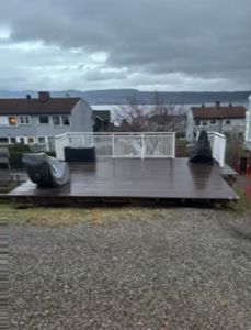 Fotografi av en terrasse med hvit hekk og dekket grill, sett fra bakgården mot en vannflate.