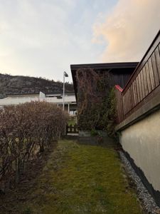 Fotografi av en bygning sett fra bakgård eller sidehage, med grøft, hekk og fjell i bakgrunnen.