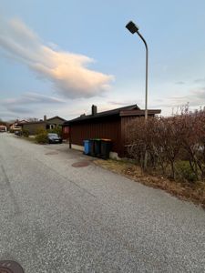 Fotografi av en eksisterende bygning (ettasjes hus med mørk fasade) sett fra gaten, med søppelcontainere og en gatelykt i forgrunnen.