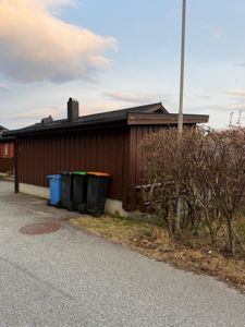 Fotografi av en brun, enetasjes bygning sett fra siden. Bildet viser fasaden, taket, en røykledning, en vei med et manhole-deksel, og en rekke søppelcontainere langs husveggen.
