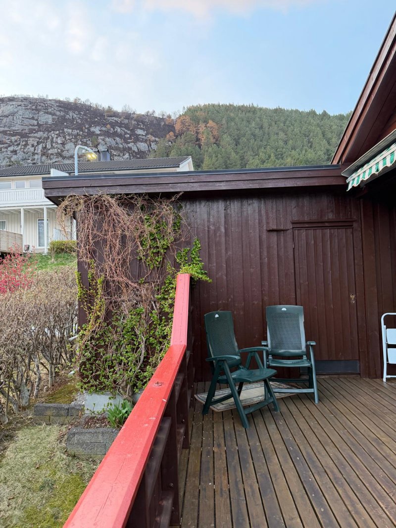 Fotografi av en terrasse med to grønne stoler, en mørk trevegg med klatreplanter og en rød balustrer i forgrunnen. I bakgrunnen ser man en annen bolig og en fjellside.