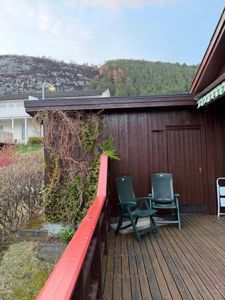 Fotografi av en terrasse med to grønne stoler, en mørk trevegg med klatreplanter og en rød balustrer i forgrunnen. I bakgrunnen ser man en annen bolig og en fjellside.