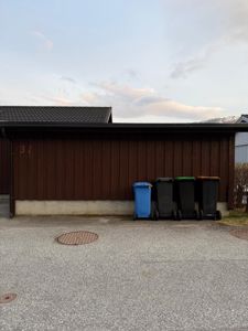 Fotografi av en brun garasje eller kjellervegg med husnummer 31, med søppelcontainere foran og en asfaltert overflate.