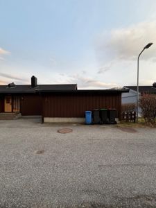 Fotografi av en enebolig sett fra foran, med synlig fasade, inngang, søppelcontainere og fortau.