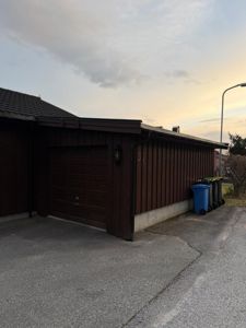 Fotografi av en mørk garasje eller uthus med tak, garasjeport og søppelcontainere foran.