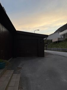 Fotografi av en mørk garasje med en betongkjørevei foran, sett mot en skråning med andre boliger.