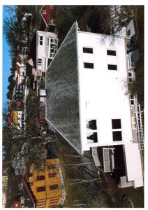 Fotografi av en hvit bygning sett fra luft eller høyde, med omgivende hus og natur.