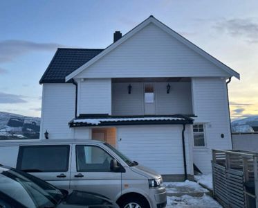 Fotografi av en hvit, toetasjes bolig med mørk takflate, sett fra foran med bil og snø i forgrunnen.