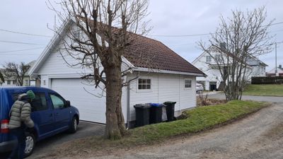 Fotografi av en hvit garasje med rød takflis, sett fra siden. Bildet viser også en blå bil, en person, og nabolaget.