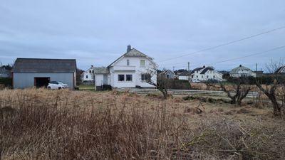 Fotografi av en hvit husfront med et grått skur til venstre, sett fra et felt.