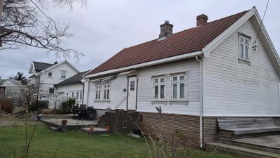 Fotografi av en hvit, trepanelt enebolig sett fra siden. Bildet viser fasaden, taket, vinduer, dør, samt deler av hagen og nabohus i bakgrunnen.