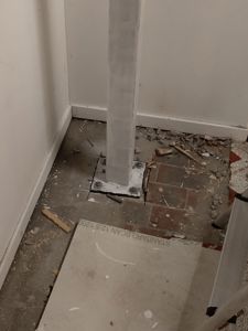 Fotografi av en byggeplass eller renovering som viser en betongstolpe festet til gulvet med bolter, omgitt av bruddstein og et gulvplater.