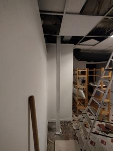 Fotografi av et rom under renovering eller ombygging, med synlig delvis demontert tak, en stolpe, trappetrinn og byggemateriale.