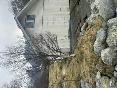 Fotografi av en hvit, panelt husfasade sett fra siden, med en liten bjørketrær foran og en steinmur til høyre.