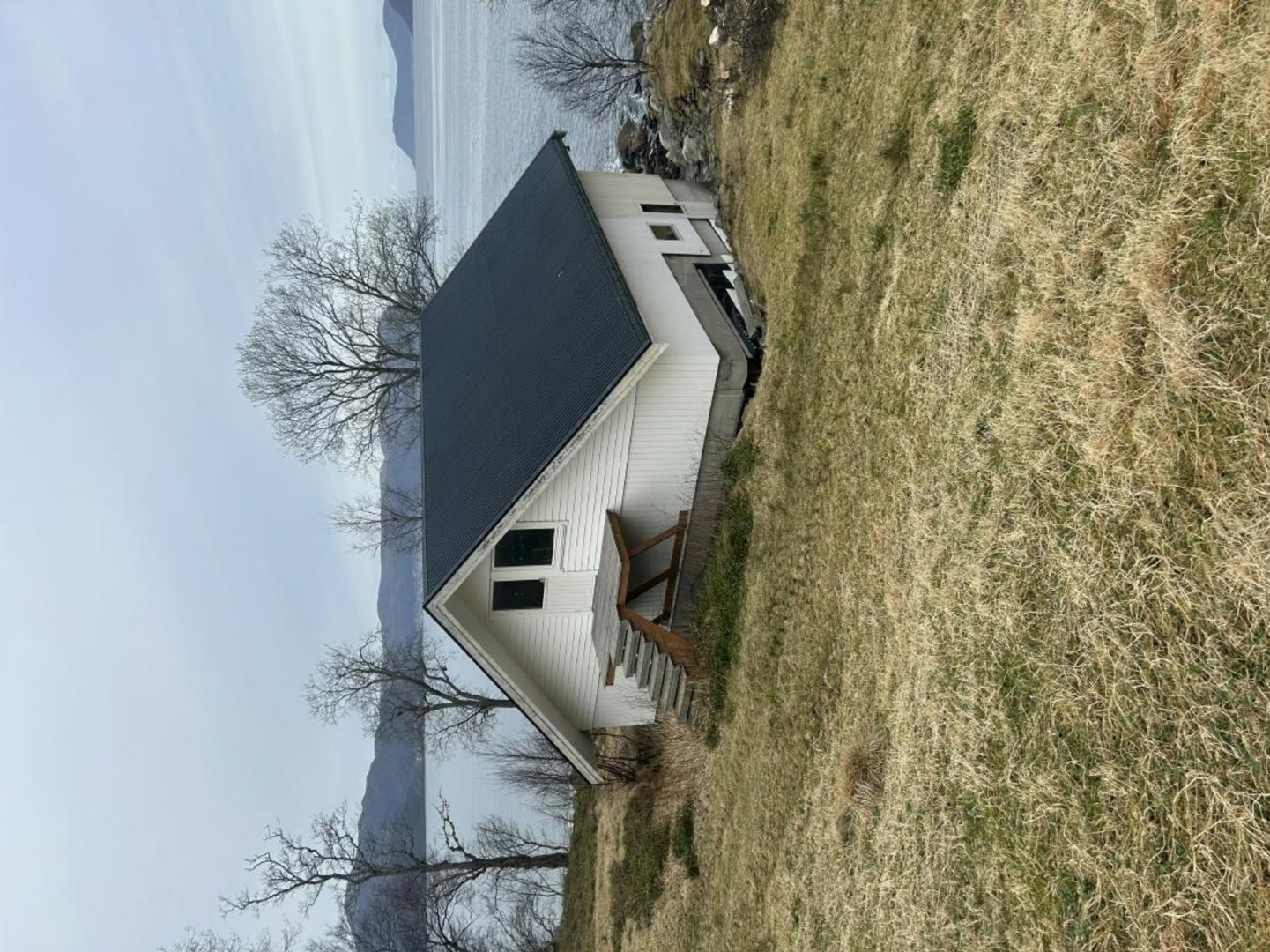 Fotografi av en liten, hvit hytte med mørkt tak, sett fra siden. Hytta ligger på en grønt/brun grasbeite med utsikt mot vann og fjell i bakgrunnen.