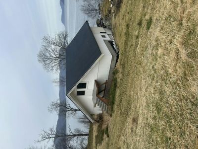 Fotografi av en liten, hvit hytte med mørkt tak, sett fra siden. Hytta ligger på en grønt/brun grasbeite med utsikt mot vann og fjell i bakgrunnen.