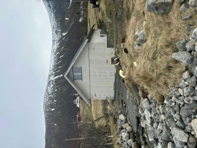 Fotografi av en liten, hvit hytte eller uthus plassert på en bratt, steinete bakke med snødekte fjell i bakgrunnen.