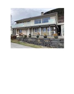 Fotografi av en toetasjes bolig sett fra foran, med en steinmur i forgrunnen og en bakkebakke i bakgrunnen.