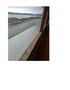 Fotografi av en balkong med glassrailing sett fra innendørs, mot et vann og fjell i bakgrunnen.