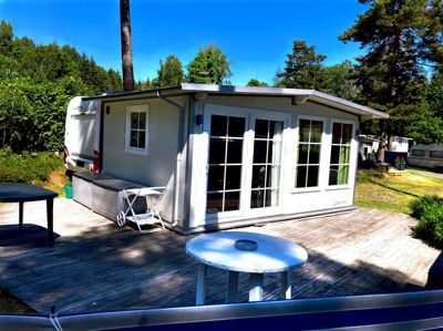 Fotografi av en hvit hytte eller campinghytte med en terrasse, plassert i en skogstrakt.