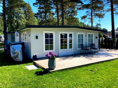 Fotografi av en hvit campinghytte med en terrasse, omgitt av grønt og trær.