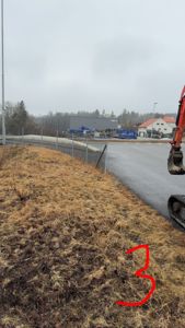 Fotografi av et bygge- eller anleggsområde med grusvei, hekk, og bygninger i bakgrunnen.