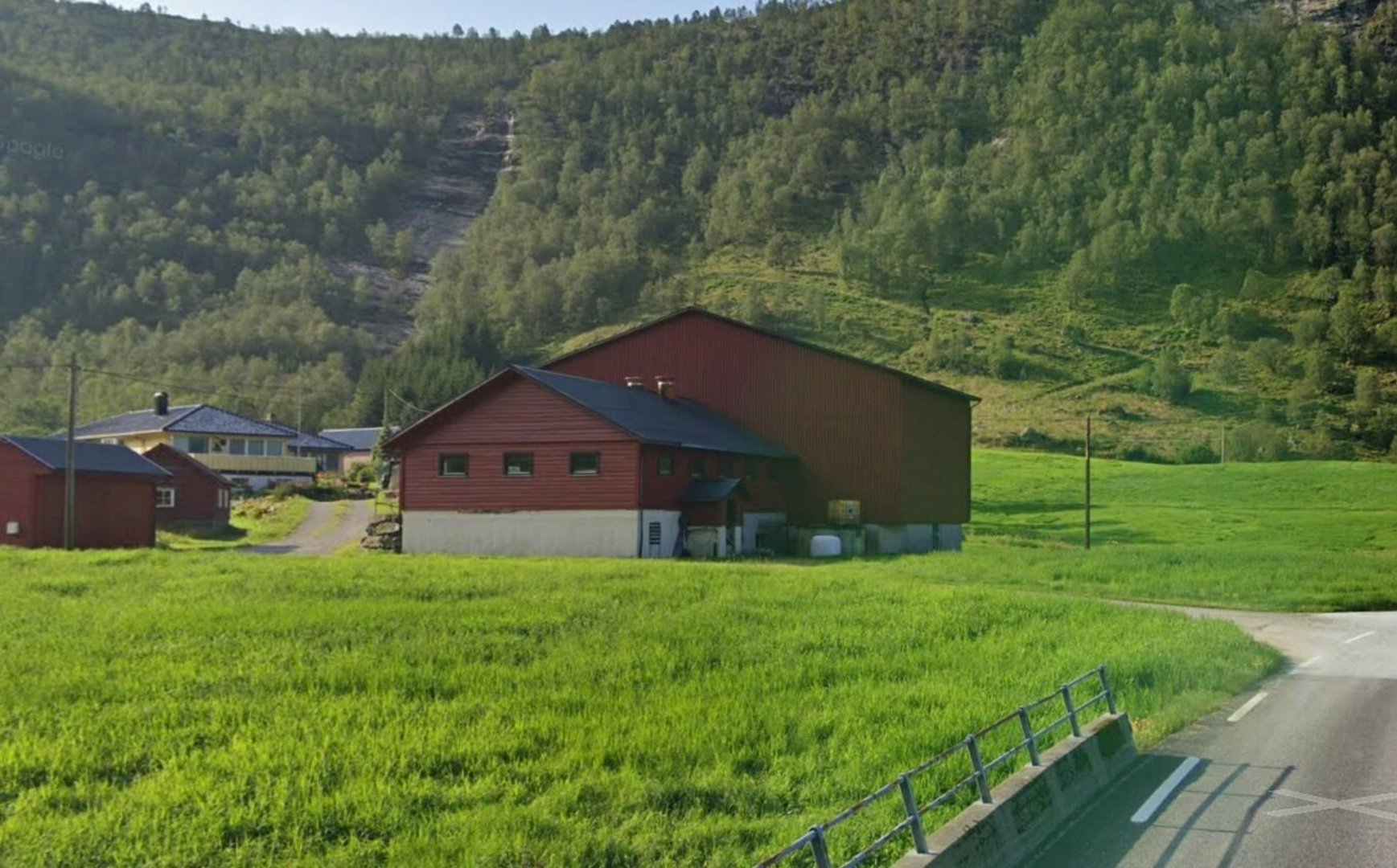 Fotografi av en rød bygning (sannsynligvis en uthus eller garasje) i en grønn, landlig omgivelser med fjell i bakgrunnen.