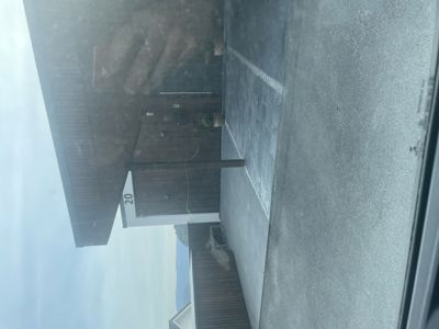 Fotografi av en bygning sett fra utsiden, med en betongplass foran og en del av en vei eller parkeringsplass til høyre. Bildet er tatt gjennom et vindusglass, noe som gir refleksjoner.