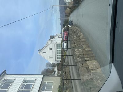 Fotografi av en hvit, trepanelt bygning sett fra en vei, med bilparkering og en steinmur i forgrunnen.