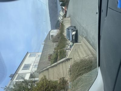 Fotografi tatt fra en bil som viser en vei med trapper, en bil parkert, og en hvit bygning i bakgrunnen.