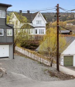 Fotografi av en boligbebyggelse med flere hus, garasjer, trær og en vei i et skrått terreng.