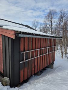 Fotografi av en rød og mørk bygning (sannsynligvis en uthus eller garasje) sett fra siden, med snø på taket og i bakken.