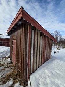 Fotografi av en liten, gammel trebygning (kanskje utedo eller skur) i snø, sett fra siden og litt forfra.