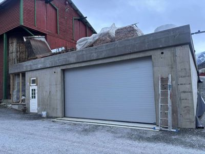 Fotografi av en nybygd betonggarasje med en stor rulleporter, en hvit dør og en trapp. I bakgrunnen står det en rød bygning.