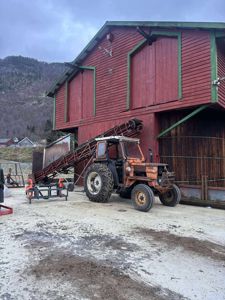 Fotografi av en rød, toetasjes bygning (muligens en uthus eller lager) med en gule traktor og en transportbåndanlegg foran. Bakgrunnen viser fjell og nabolag.