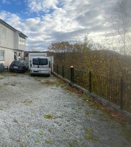 Fotografi av en grusplass foran en hvit bygning med to parkerte biler (en bil og en varebil) og en hekk i bakgrunnen.