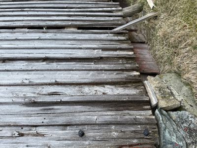 Fotografi av en gammel, utslitt treplattform eller trapp som går ned mot en steinmur og grøft.