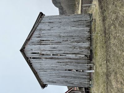 Fotografi av en gammel, forfallelig trebygning (kanskje en uthus eller skur) sett fra siden. Bygningen står på et åpent, grønt område med en fjellside i bakgrunnen.