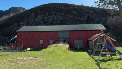 Fotografi av en stor, rød skurbygning med grønn takflate, plassert foran en steil fjellside. Bildet viser også en grønn hage med et lekeplass-utstyr i forgrunnen.
