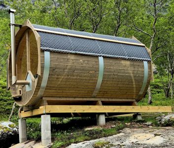 Fotografi av en ferdigbygd, sylindrisk trehytte (trollhytte) med solpaneler på taket, stående på betongfundament i et skogsmiljø.