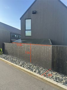 Fotografi av en mørk fasade på en bygning med en tilhørende hekk eller skillevegg. Bildet inneholder også håndtegnede målinger i rødt.