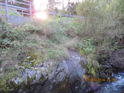 Fotografi av et natursceneri med en bro i bakgrunnen, en steinete elvebredd og vegetasjon.