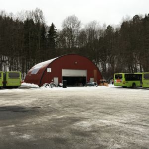 Fotografi av en rød, halvsirkelformet bygning (sannsynligvis en busstall eller verksted) med snø på bakken og busser i nærheten.