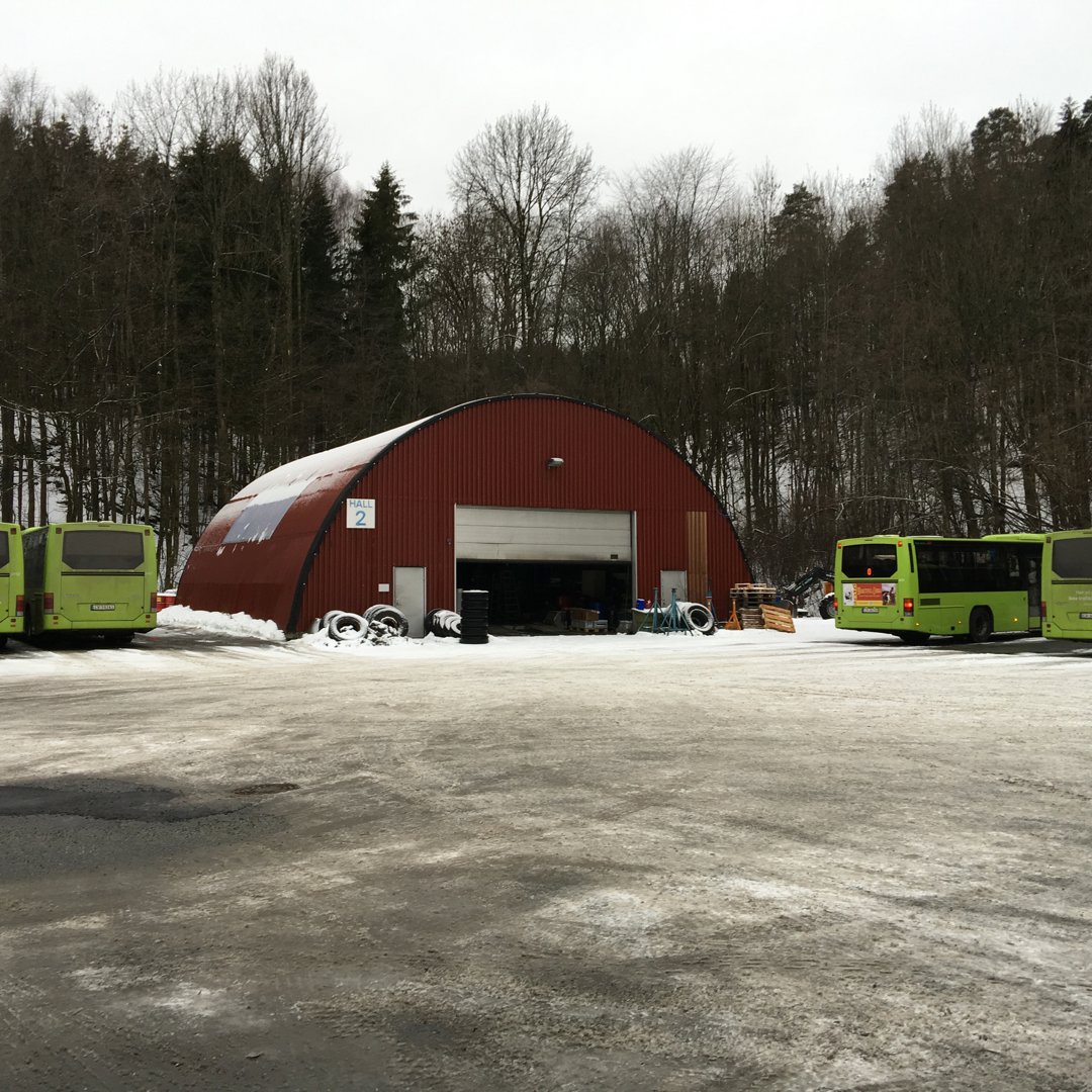 Fotografi av en rød, halvsirkulær bygning (muligens en verkstedhall eller garasje) med snø på bakken og to grønne busser parkert foran.
